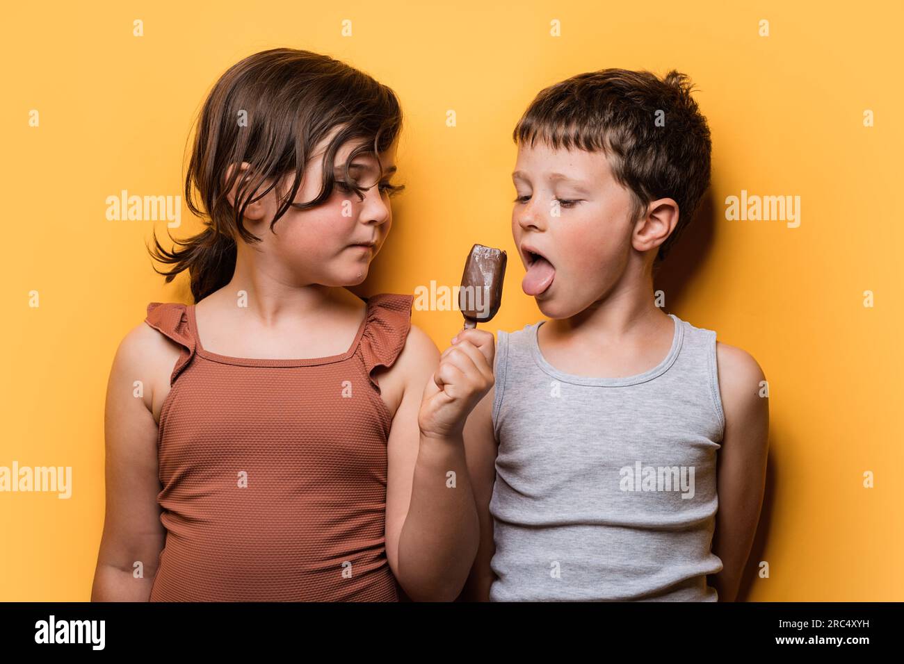 Boy licking ice cream covered in chocolate while girl in brown swimsuit sharing treat against orange background Stock Photo