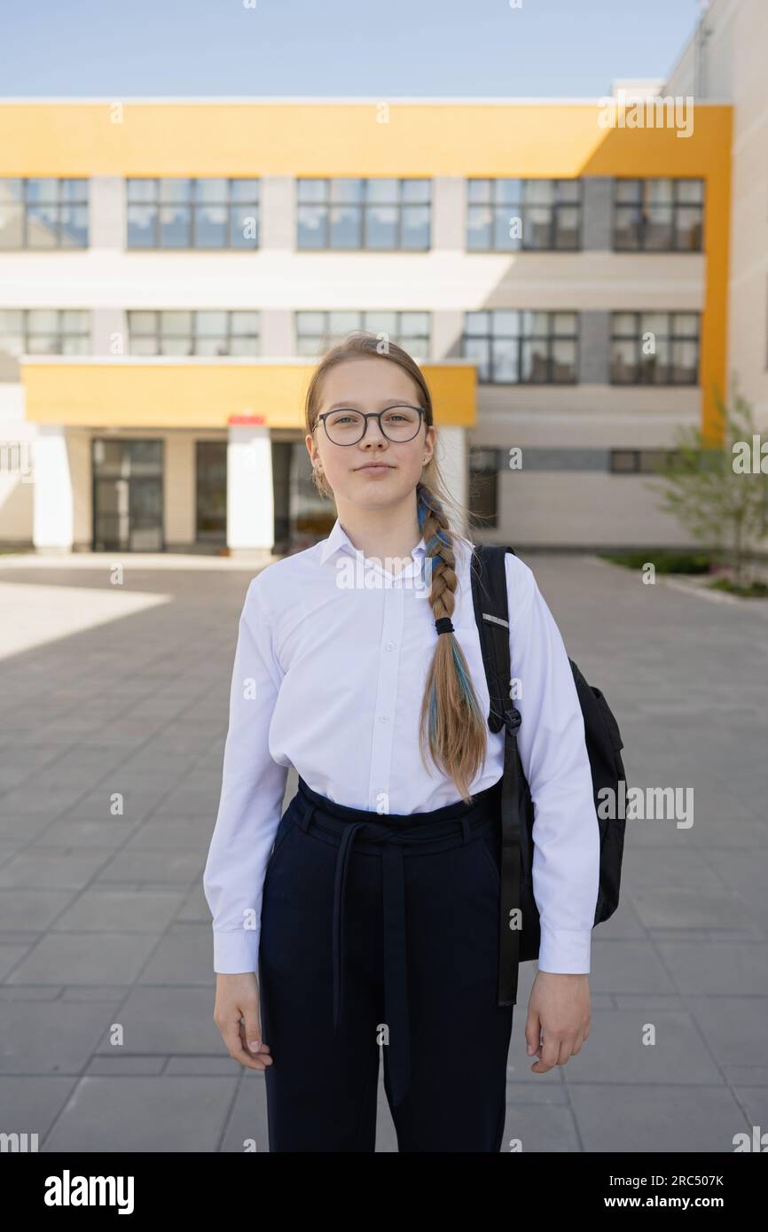 Serious young adolescent girl in uniform and with backpack looking at camera while standing on school compound near blurred glass building in daylight Stock Photo