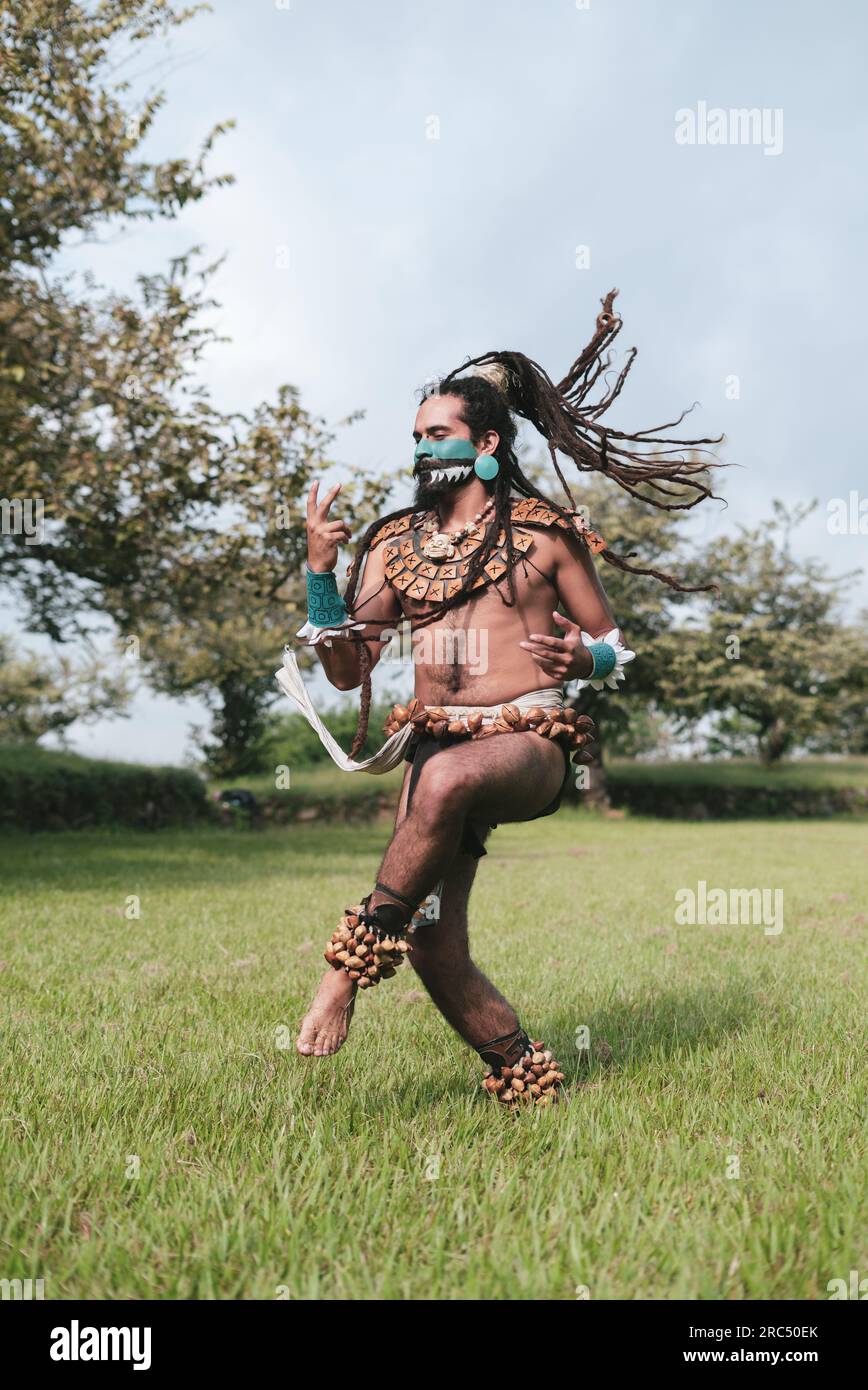 Full body of happy braided hair young male Mayan warrior in traditional costumes and body accessories dancing on grass in park at daylight Stock Photo