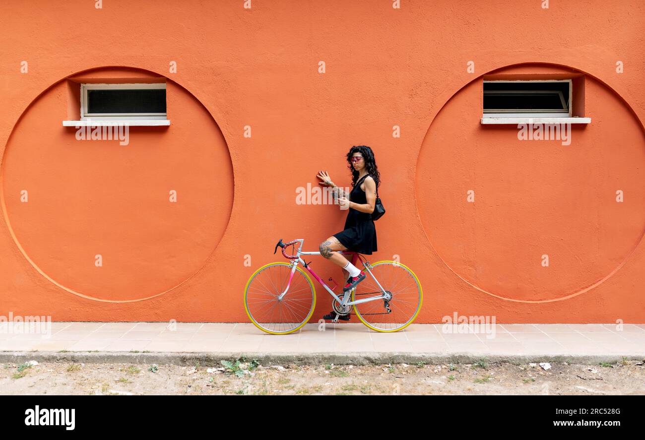 Side view of young Hispanic female looking at screen of mobile phone while sitting on cycle and holding orange wall with hand on sidewalk in daylight Stock Photo