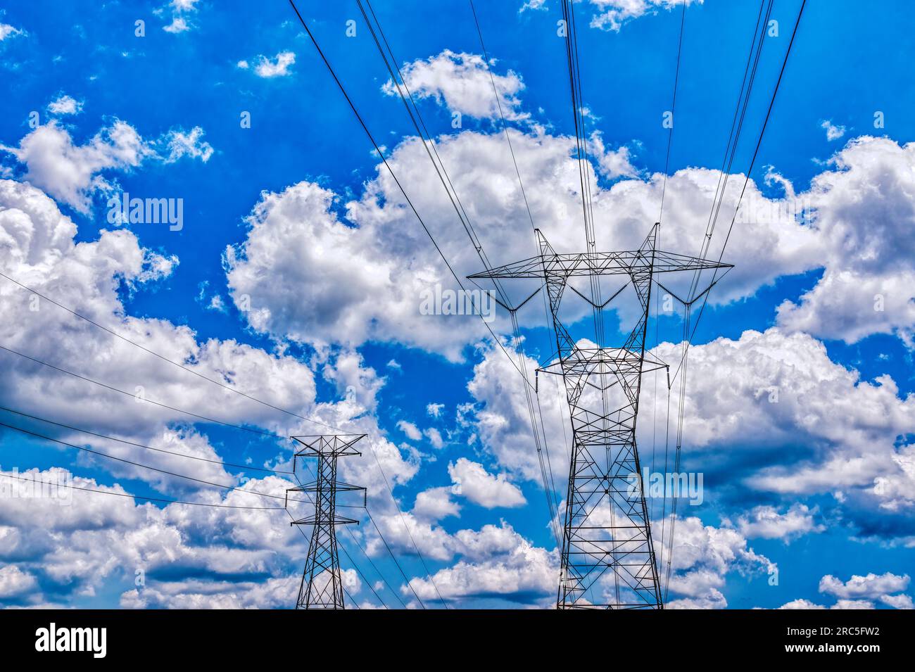 Horizontal shot of main electric towers against puffy white clouds in a blue sky. Stock Photo