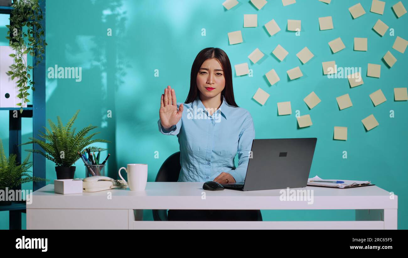 Adamant uncompromising businesswoman saying stop throwing halt sign gesture, unhappy with work conditions. Assertive asian employee at colourful office desk over blue studio background Stock Photo