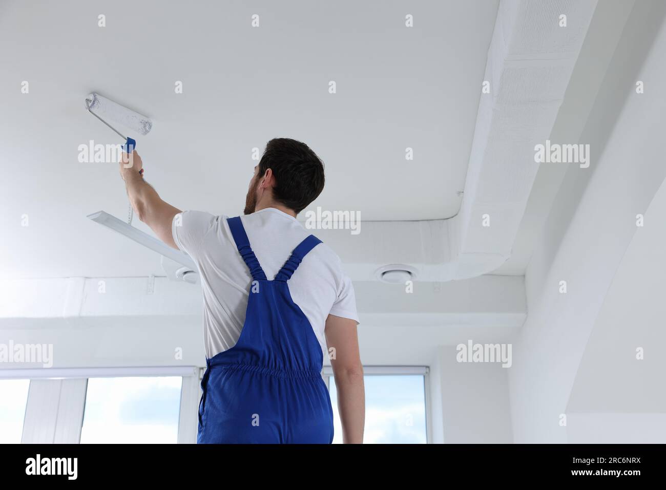 Handyman with roller painting ceiling in room, back view Stock Photo