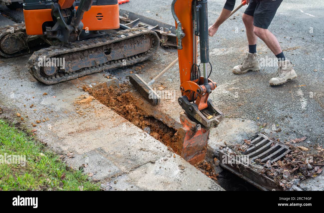 Excavator digging up dirt between cut concrete pavement. Man sweeping dirt with broom. Roadworks in Auckland. Stock Photo