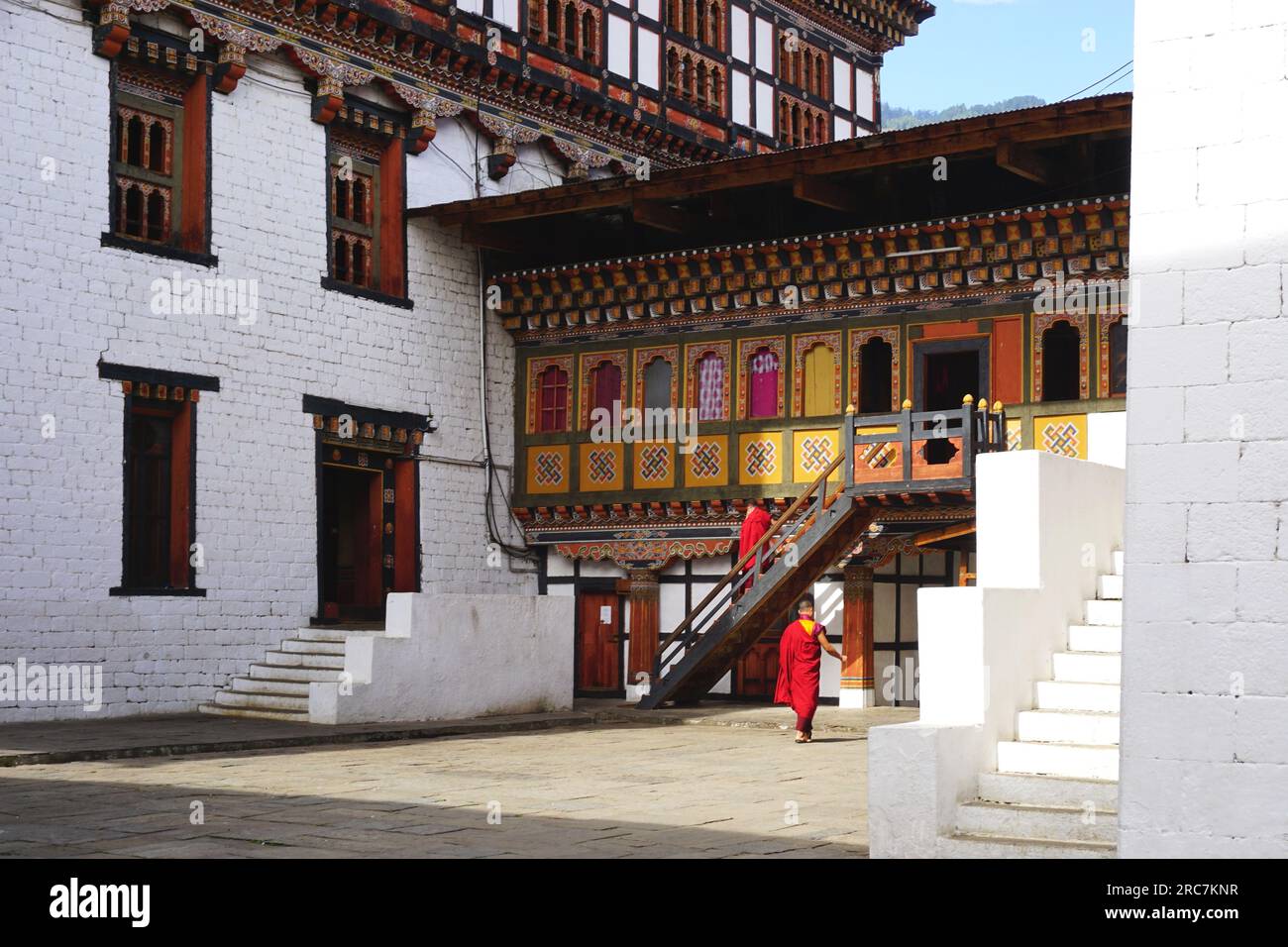 A pair of red-robed Buddhist monks walk near a traditional colorful painted building on the grounds of Tashichho Dzong in Thimphu, Bhutan. Stock Photo
