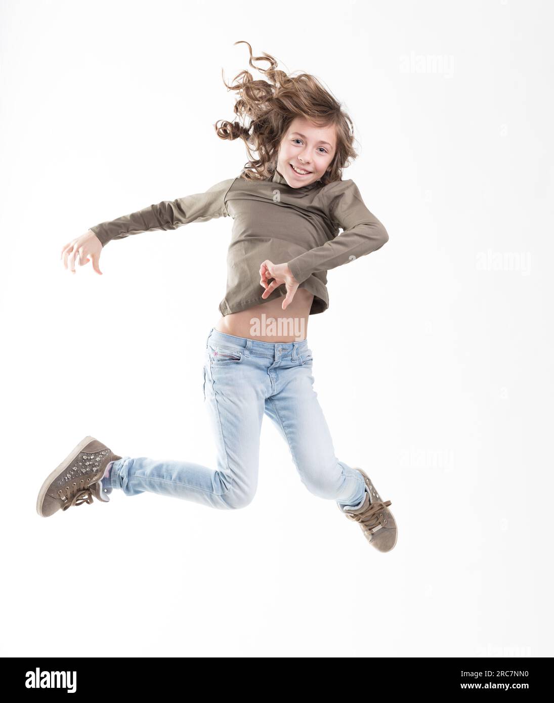 Isolated on white, a brown-curled girl in mid-leap, joy etched on her face. Dressed in blue jeans, green shirt, she loves to fly Stock Photo