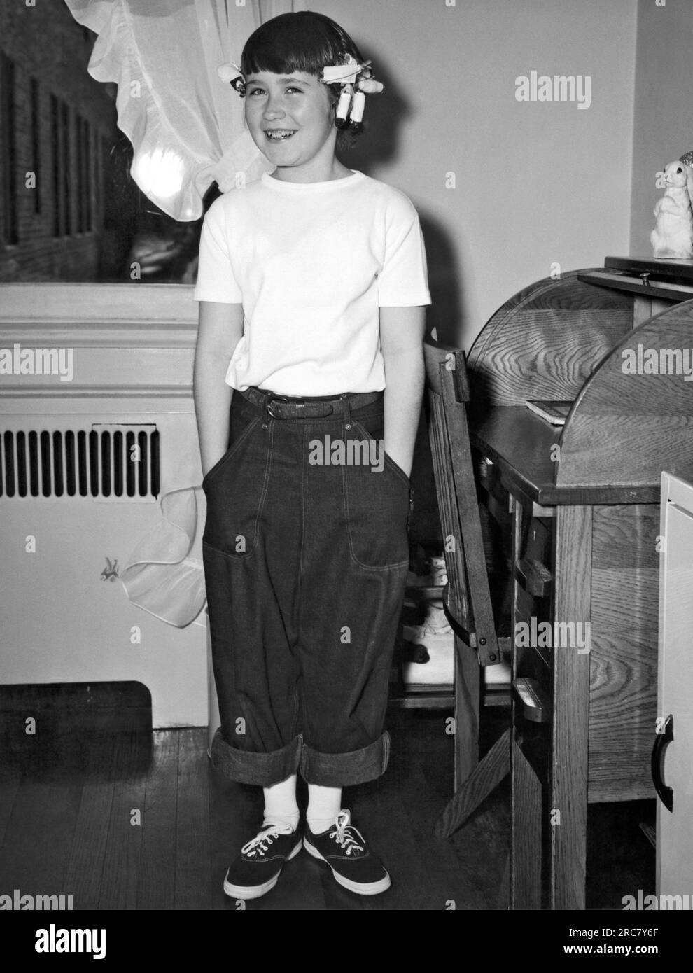New York, New York:  c. 1940 A smiling young girl wearing rolled up jeans, tennis shoes, and curlers in her hair stands by her desk in her room. Stock Photo