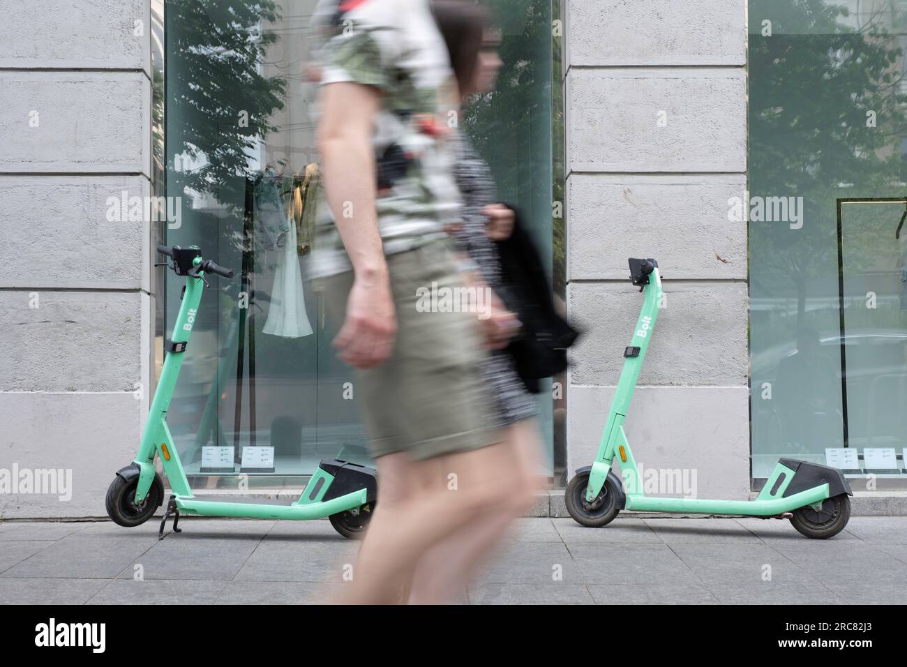 Shared electric kick scooters for rent in front of a fashion store window. A couple walks by (motion blur). E-mobility Stock Photo