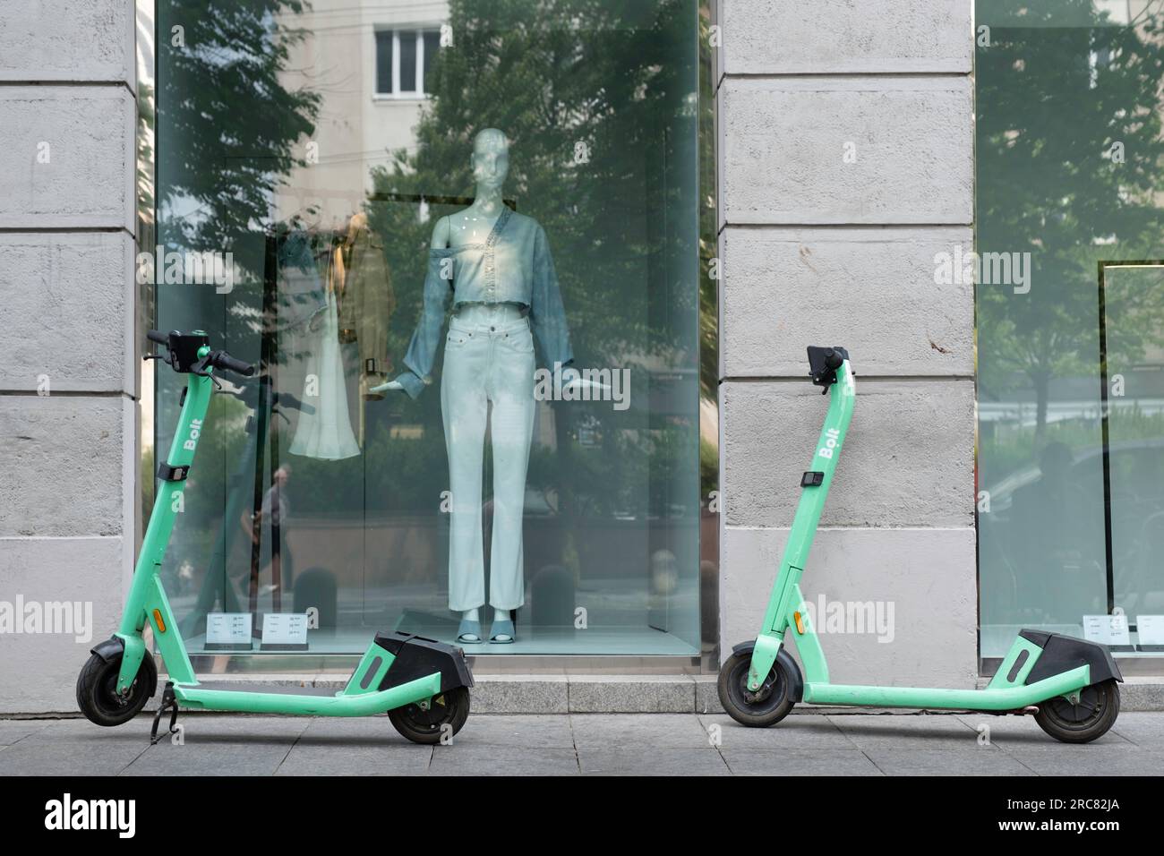Shared electric kick scooters for rent in front of a fashion store window. A model in the window watches and gestures with her hands. E-mobility Stock Photo