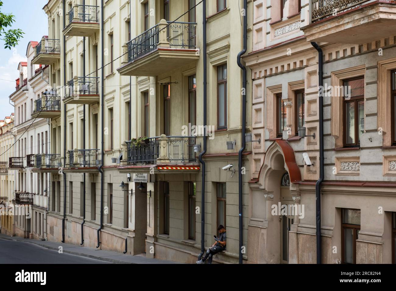 Descending street in Vilnius with facades of old gloomy historical buildings in pastel shades, many with balconies. A man looks at his phone Stock Photo
