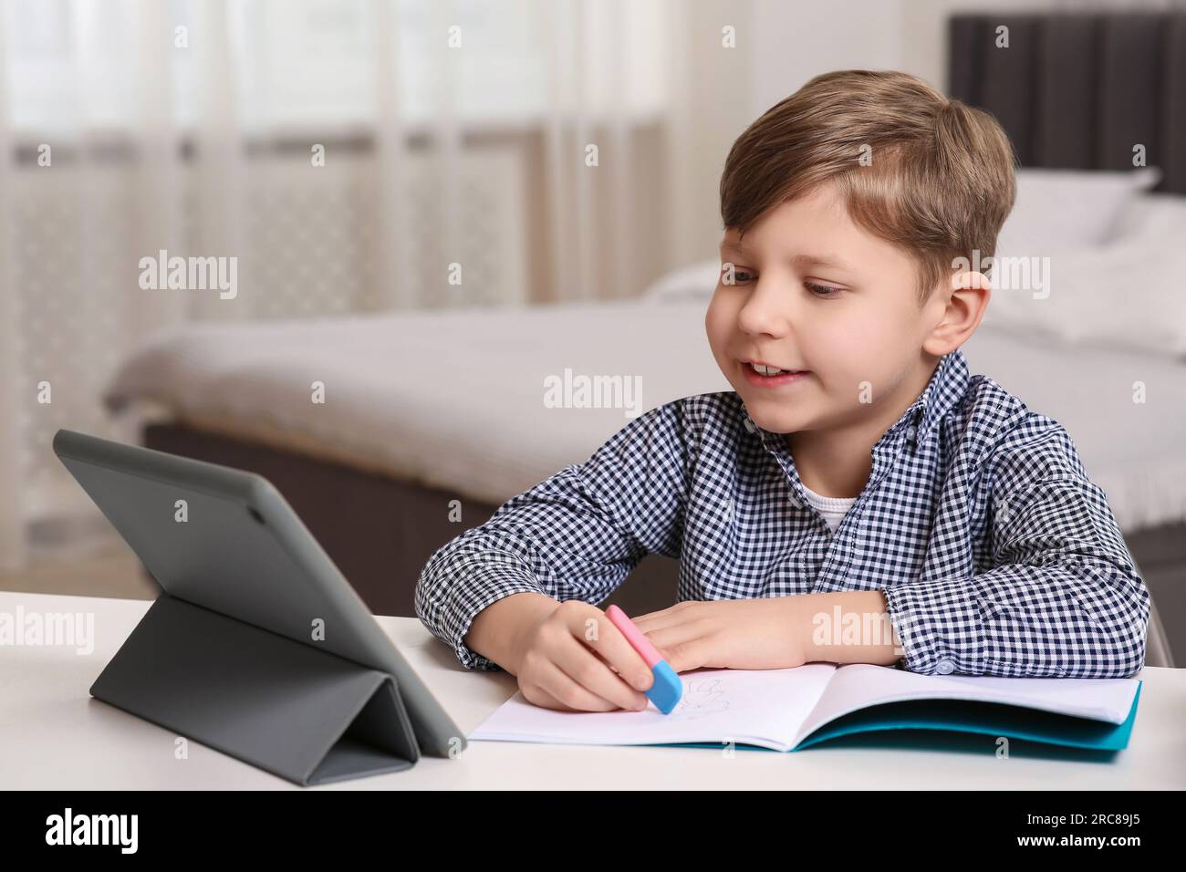 Little boy erasing mistake in his notebook near tablet at white desk indoors Stock Photo