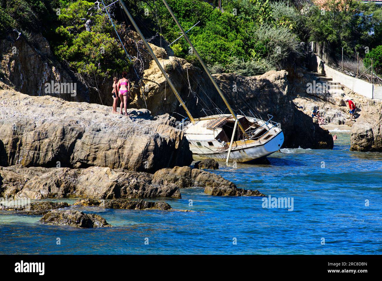 People looking at stranded boat on rocky coastline, surrounded by nature's beauty and serene waters. Stock Photo