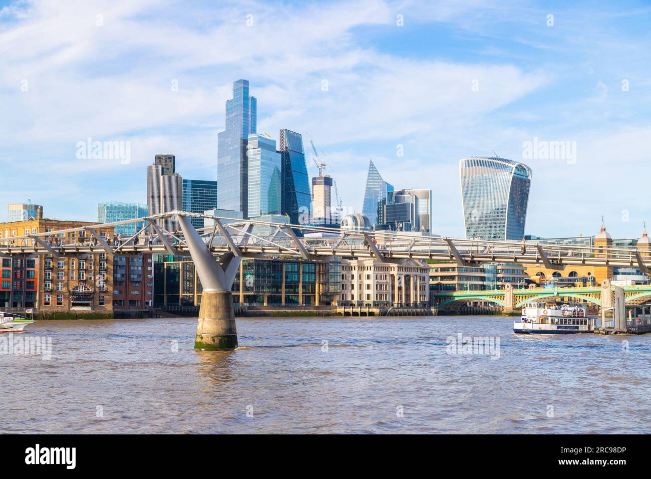 LONDON, UK - 6TH JULY 2023: The Millennium Bridge and City of London skyline during the day. People can be seen on a boat and the bridge. Stock Photo