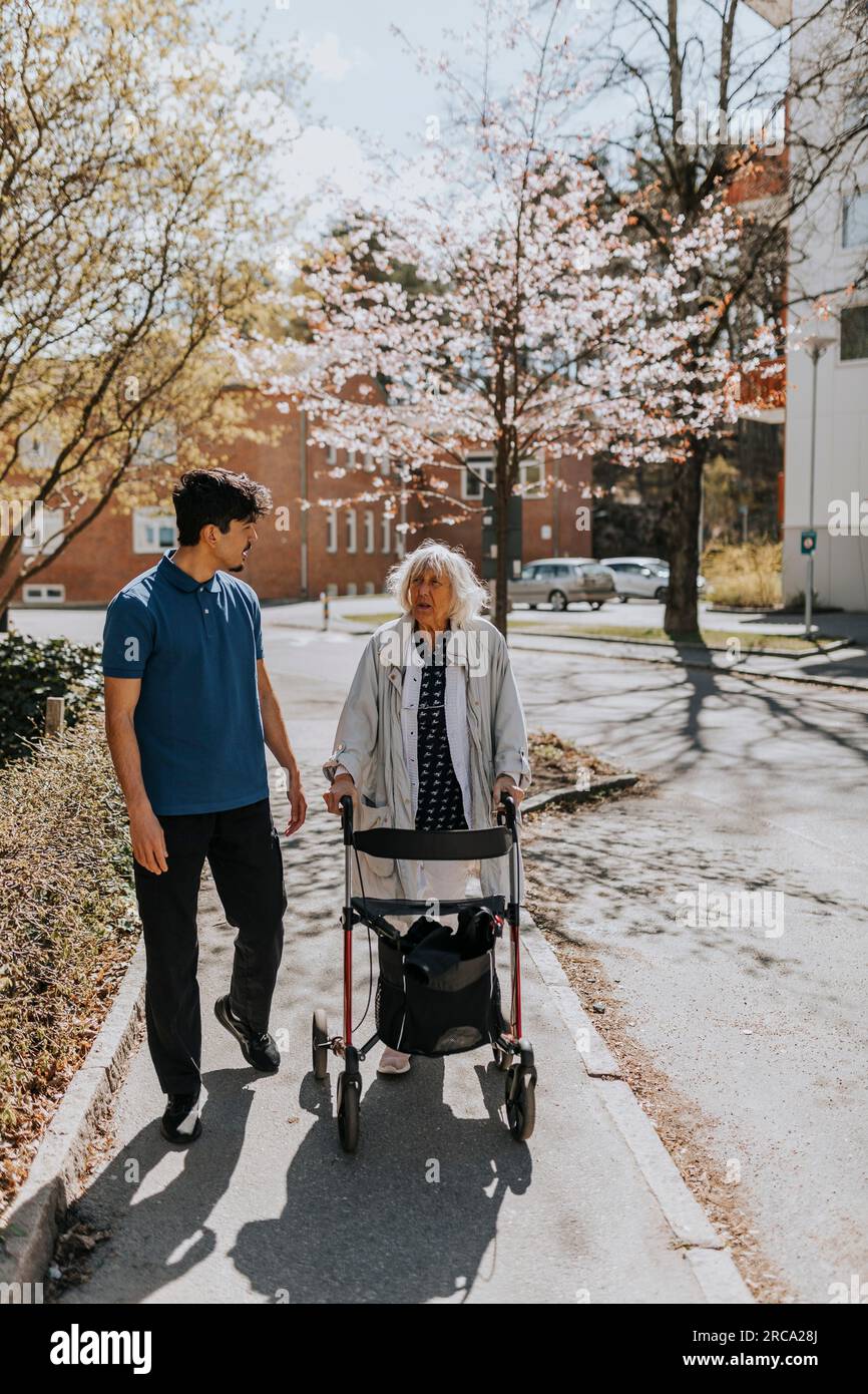 Male caregiver walking with senior woman using walker on sidewalk Stock Photo