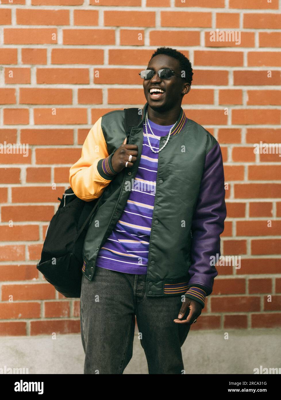 Happy young man with backpack while standing in front of brick wall Stock Photo