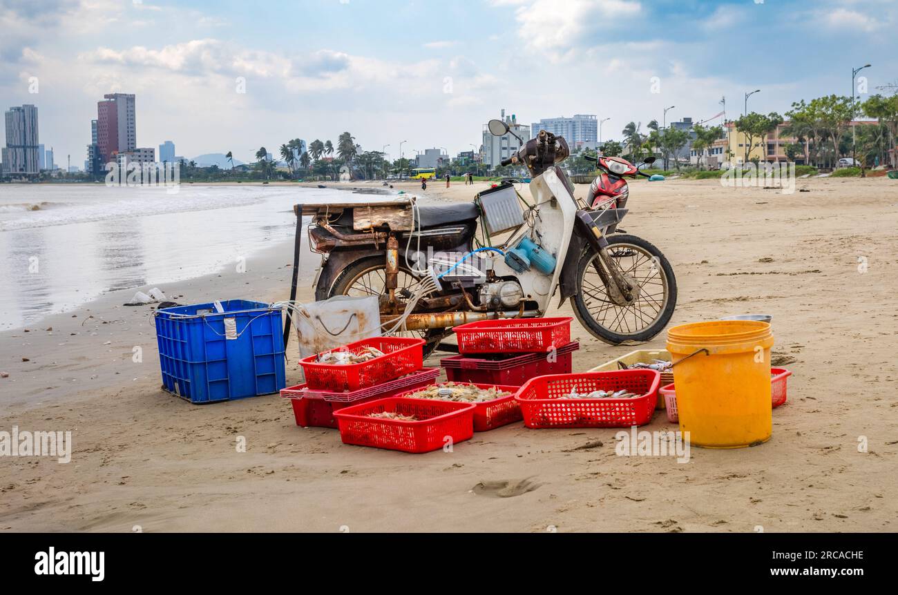 An old and rusty scooter carrying a car battery and next to trays of fish and seafood on My Khe beach in Son Tra, Danang, Vietnam. Stock Photo