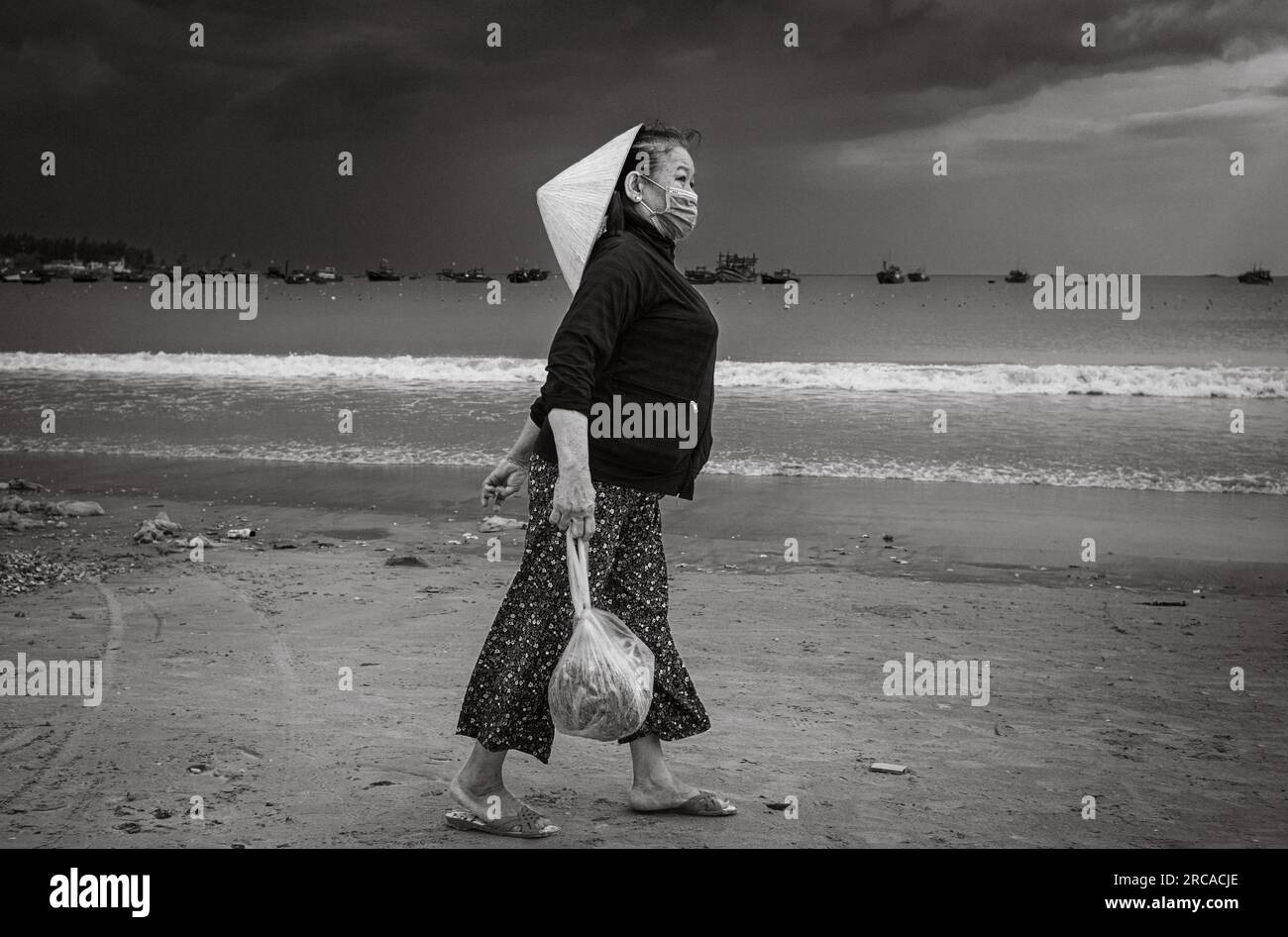 A middle-aged Vietnamese woman wearing a traditional conical hat carries a bag of seafood on My Khe beach, Danang, Vietnam. Stock Photo