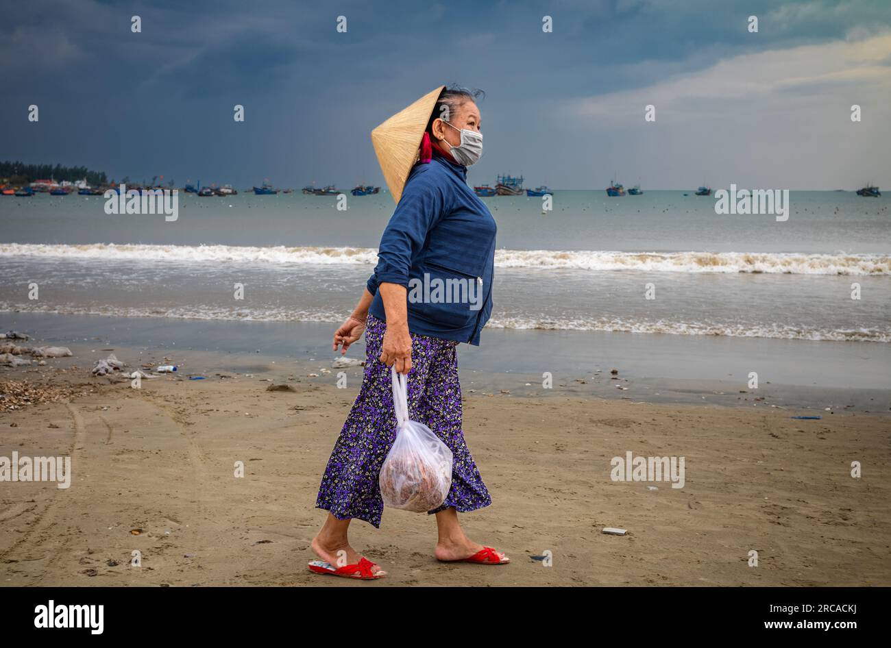 A middle-aged Vietnamese woman wearing a traditional conical hat carries a bag of seafood on My Khe beach, Danang, Vietnam. Stock Photo