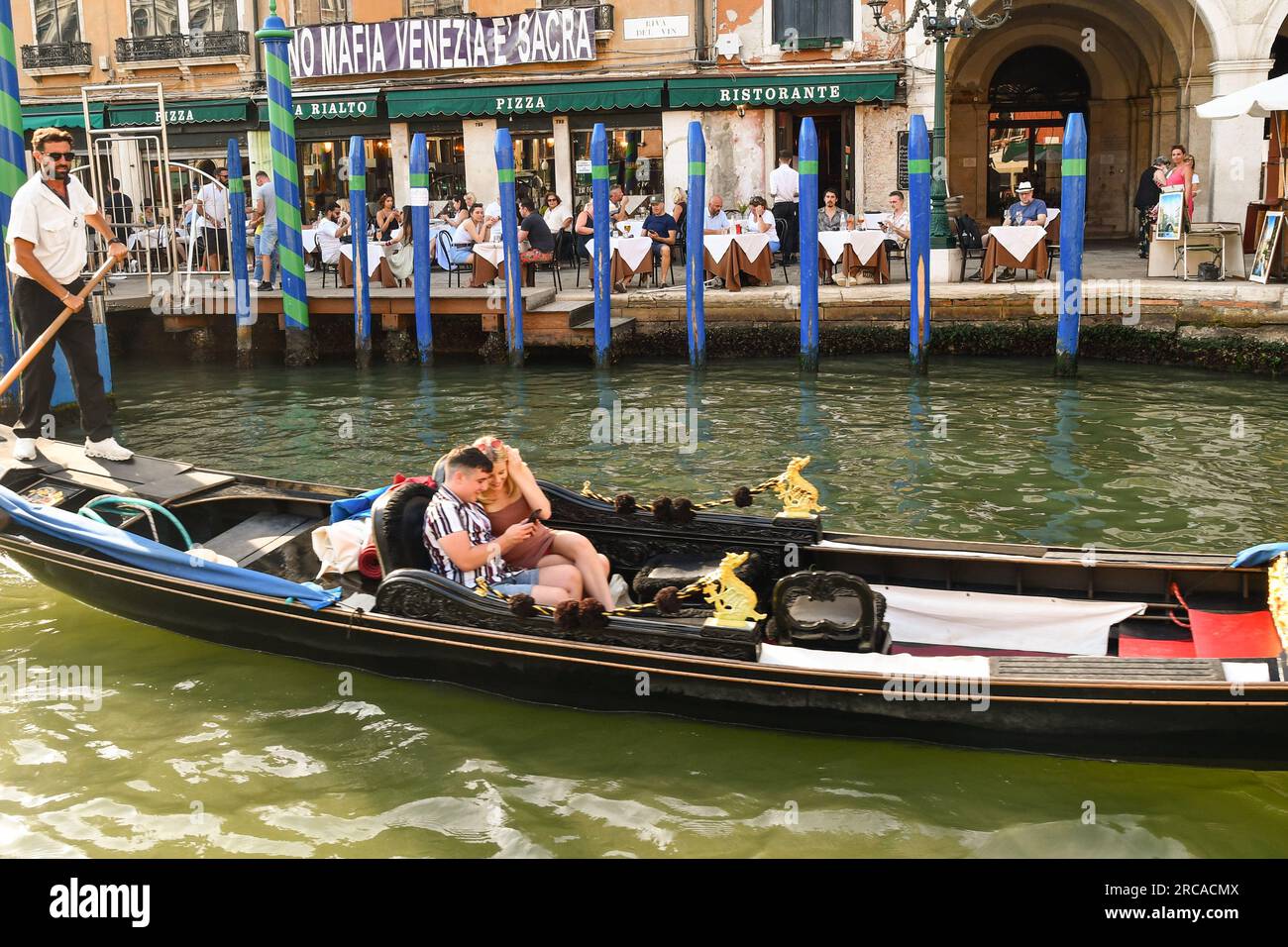 A young couple enjoying their gondola ride on the Grand Canal with a banner against the mafia in Venice on a palace on Riva del Vin waterfront, Italy Stock Photo