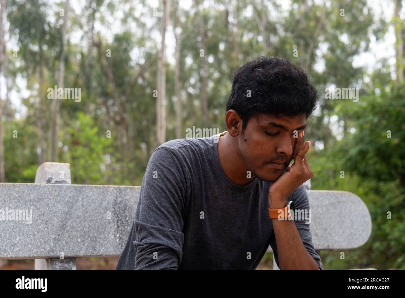 A depressed Indian man sits on a bench in a park, tears streaming down his face. He looks lost and alone, his head in his hands. Stock Photo