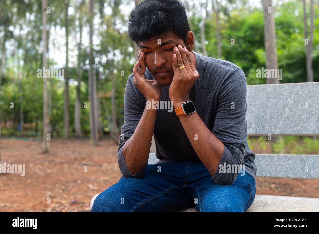 A depressed Indian man sits on a bench in a park, tears streaming down his face. He looks lost and alone, his head in his hands. Stock Photo