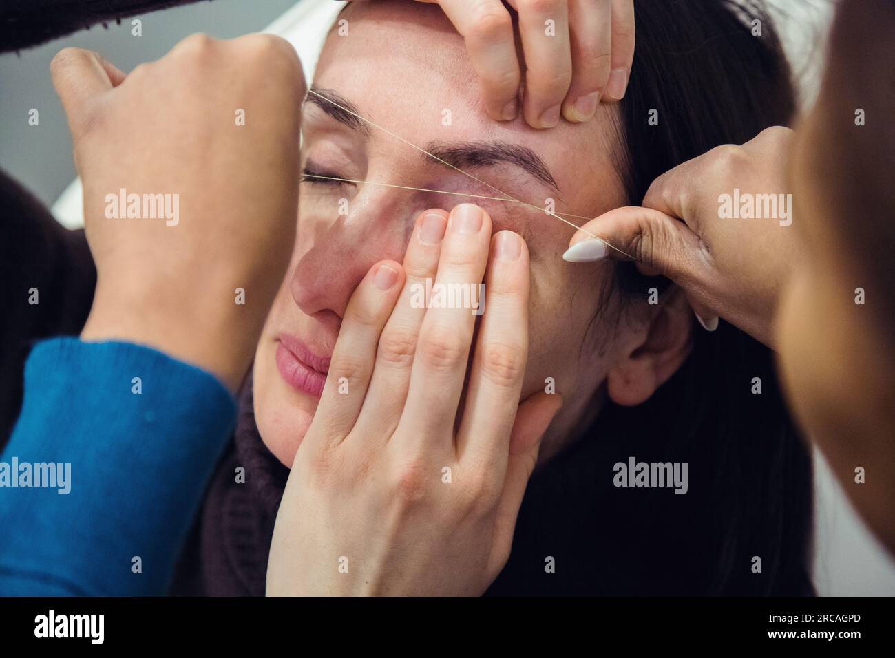 Caucasian woman removing hair from eyebrows with thread technique done by eyebrow stylist. Threading. Process Steps. Close up Stock Photo