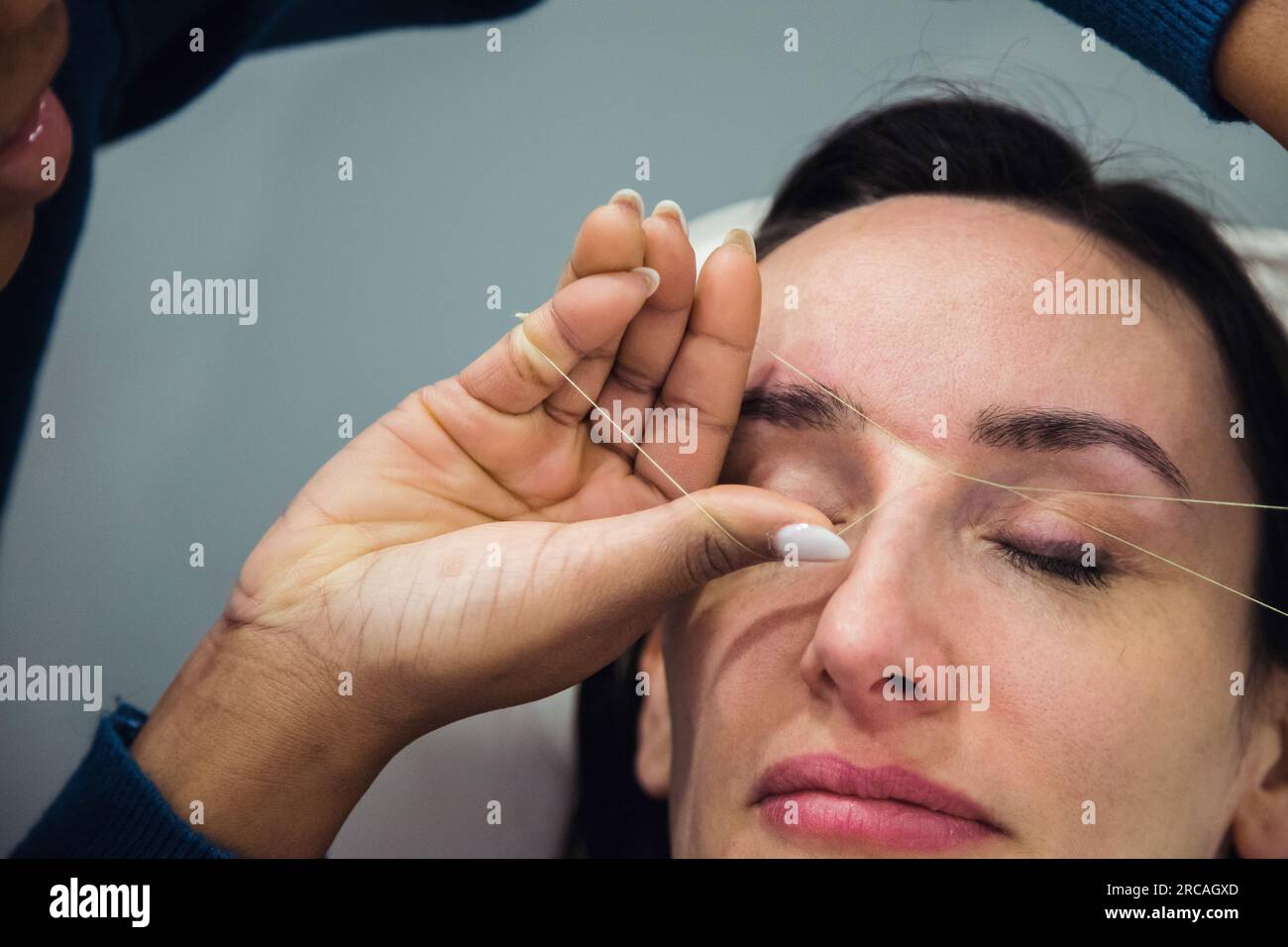Caucasian woman removing hair from eyebrows with thread technique done by eyebrow stylist. Threading. Process Steps. Close up Stock Photo