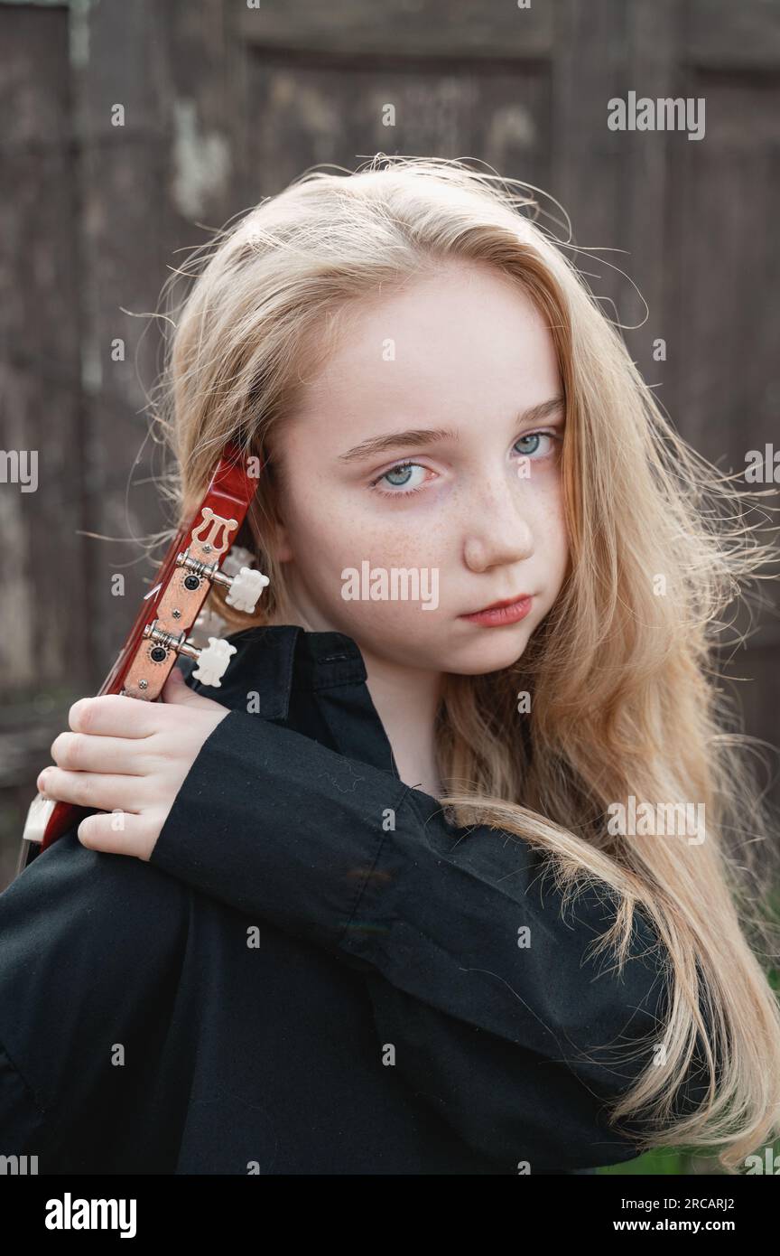 A 13-year-old teenager with a guitar. Music and hobbies. A young musician girl with long blonde hair thoughtfully holds a guitar. Sad romantic girl, f Stock Photo