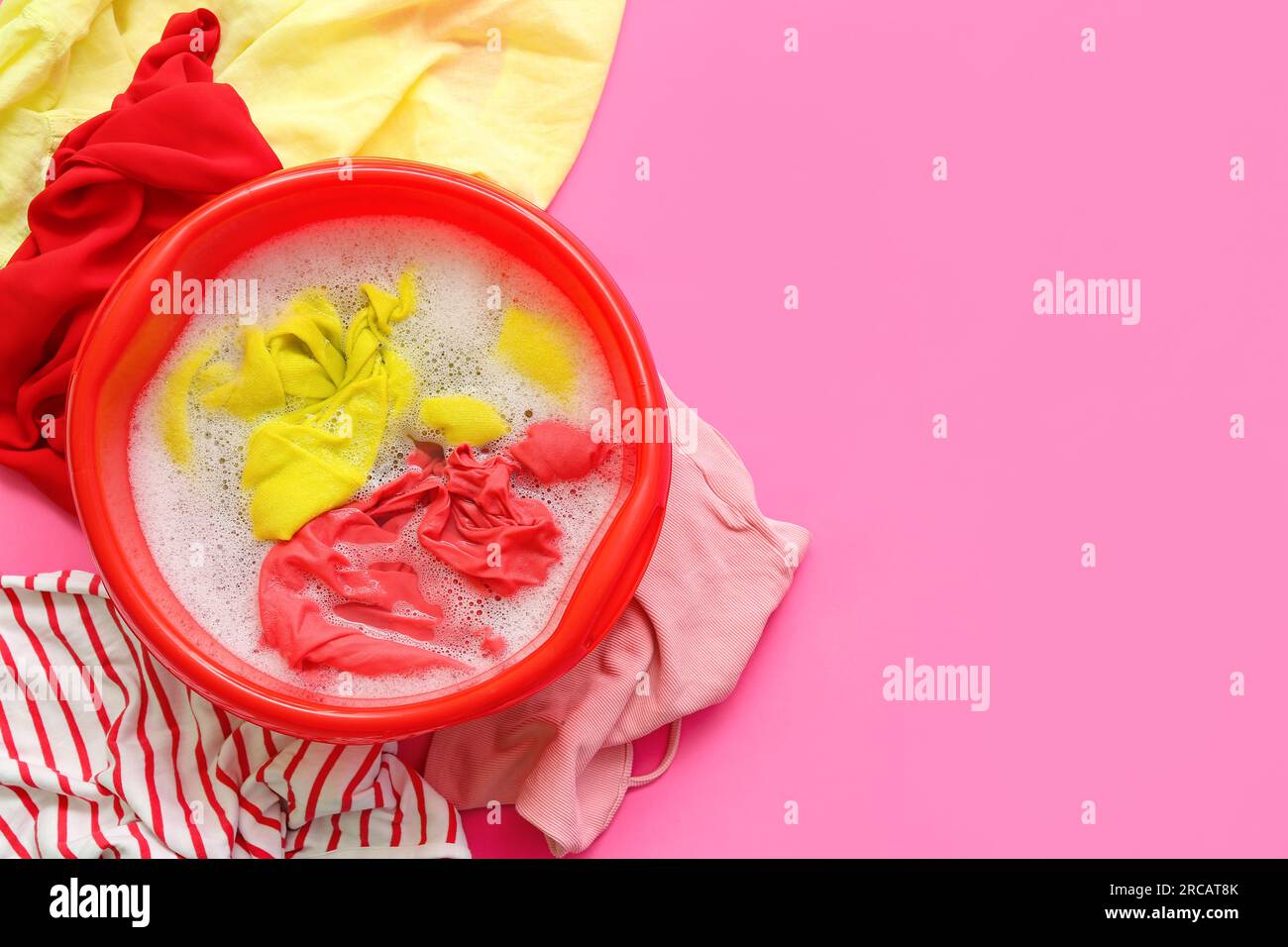 Plastic basin with dirty laundry, water and foam on pink background