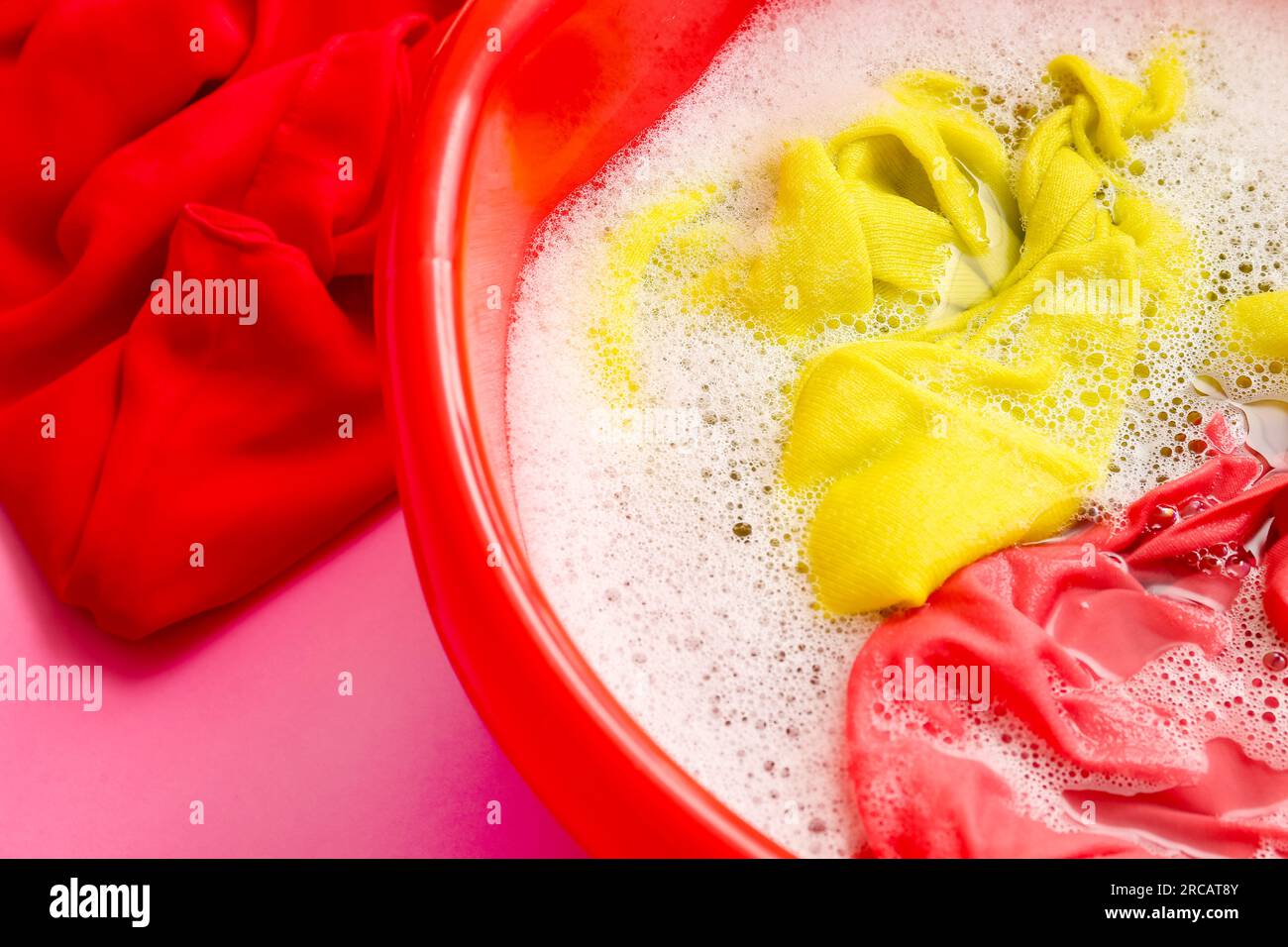Plastic basin with dirty laundry, water and foam on pink background