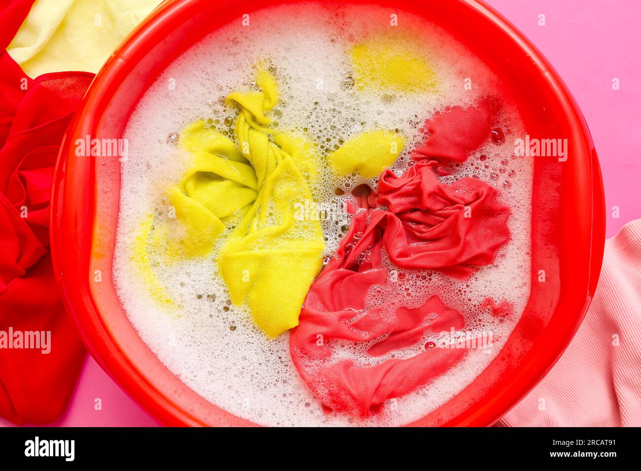 Plastic basin with dirty laundry, water and foam on pink background