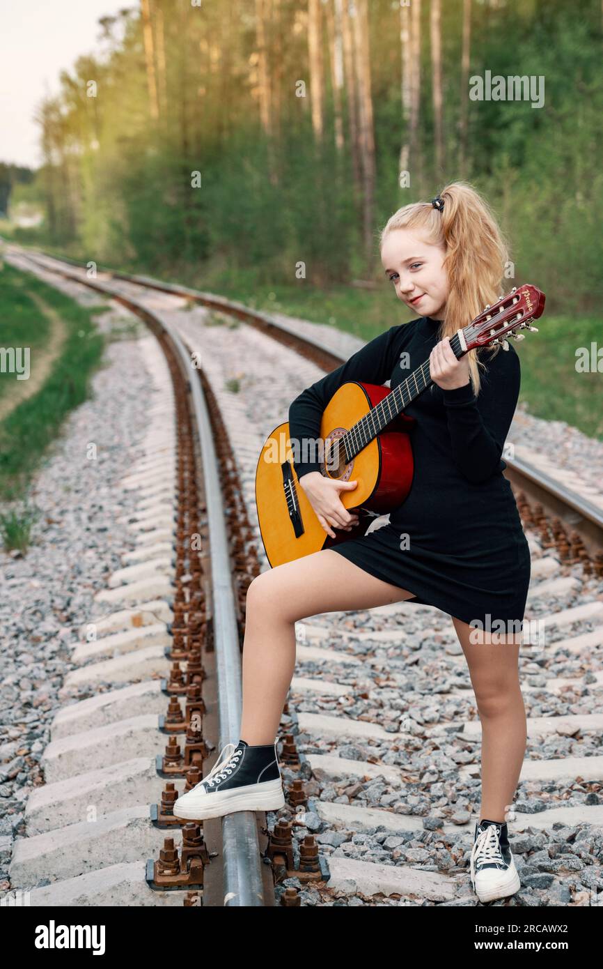 A 13-year-old teenager with a guitar. The girl travels with a guitar. The girl is standing on the rails going into the distance, in the forest at suns Stock Photo
