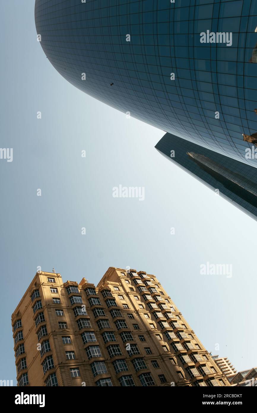 Baku, Azerbaijan - June 26, 2023: A cylindrical skyscraper's mirrored surface reflects a historic building in Baku, under a clear morning sky Stock Photo