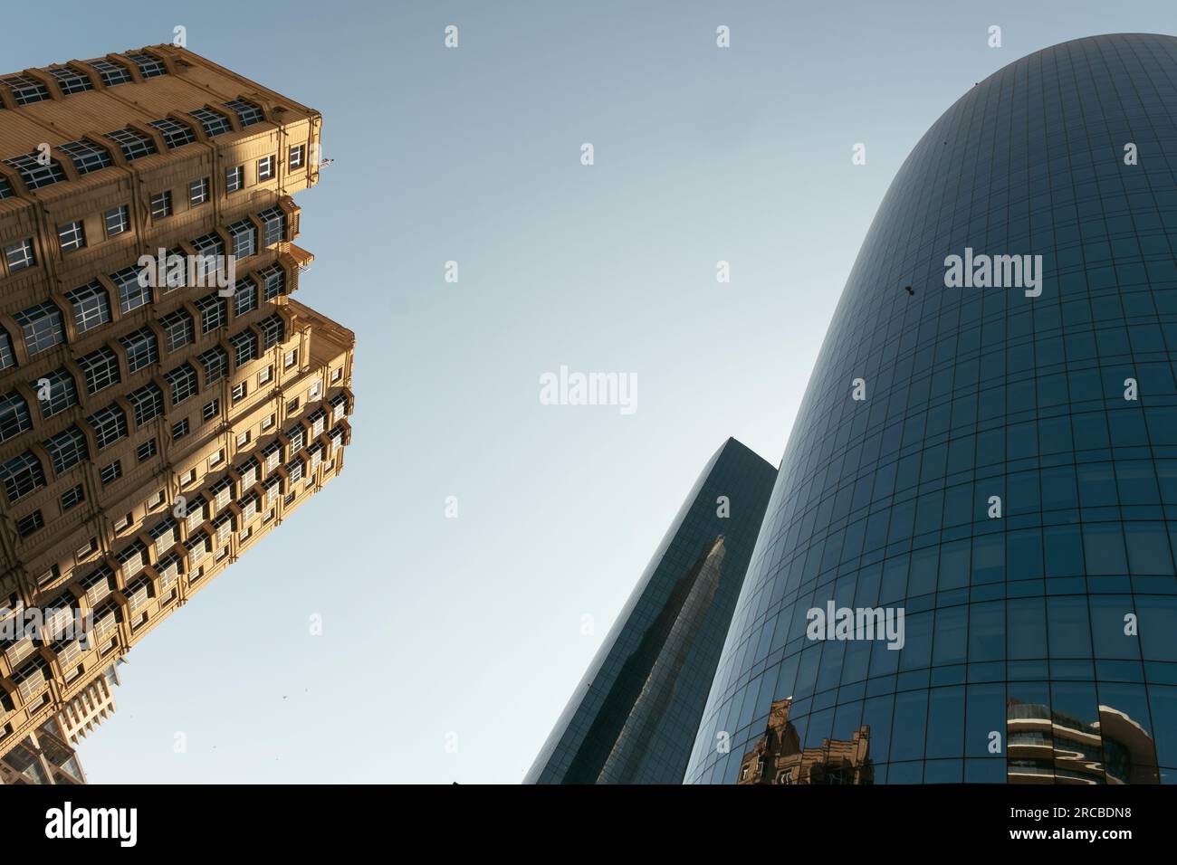Baku, Azerbaijan - June 26, 2023: A cylindrical skyscraper's mirrored surface reflects a historic building in Baku, under a clear morning sky Stock Photo