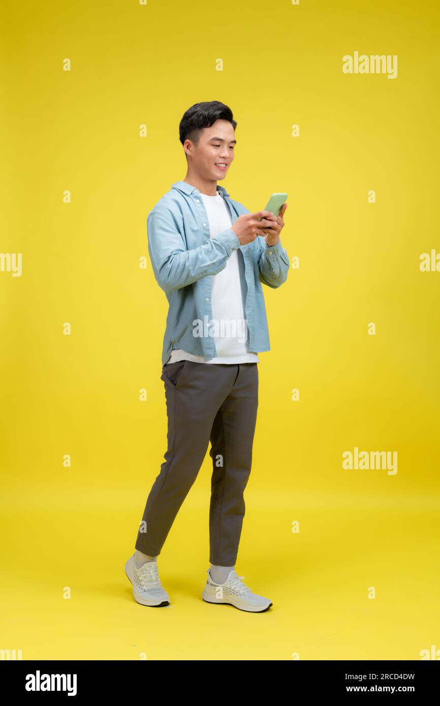 Happy man in casual wear using mobile phone chatting online, walking on yellow background Stock Photo