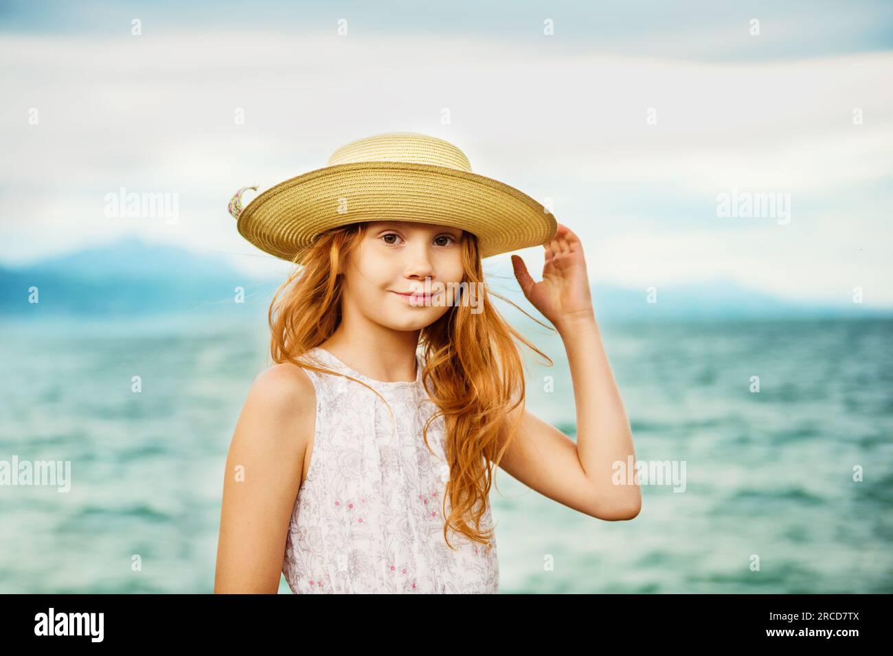 Pretty little girl with long red hair playing by the lake on a very windy day, wearing big hat Stock Photo