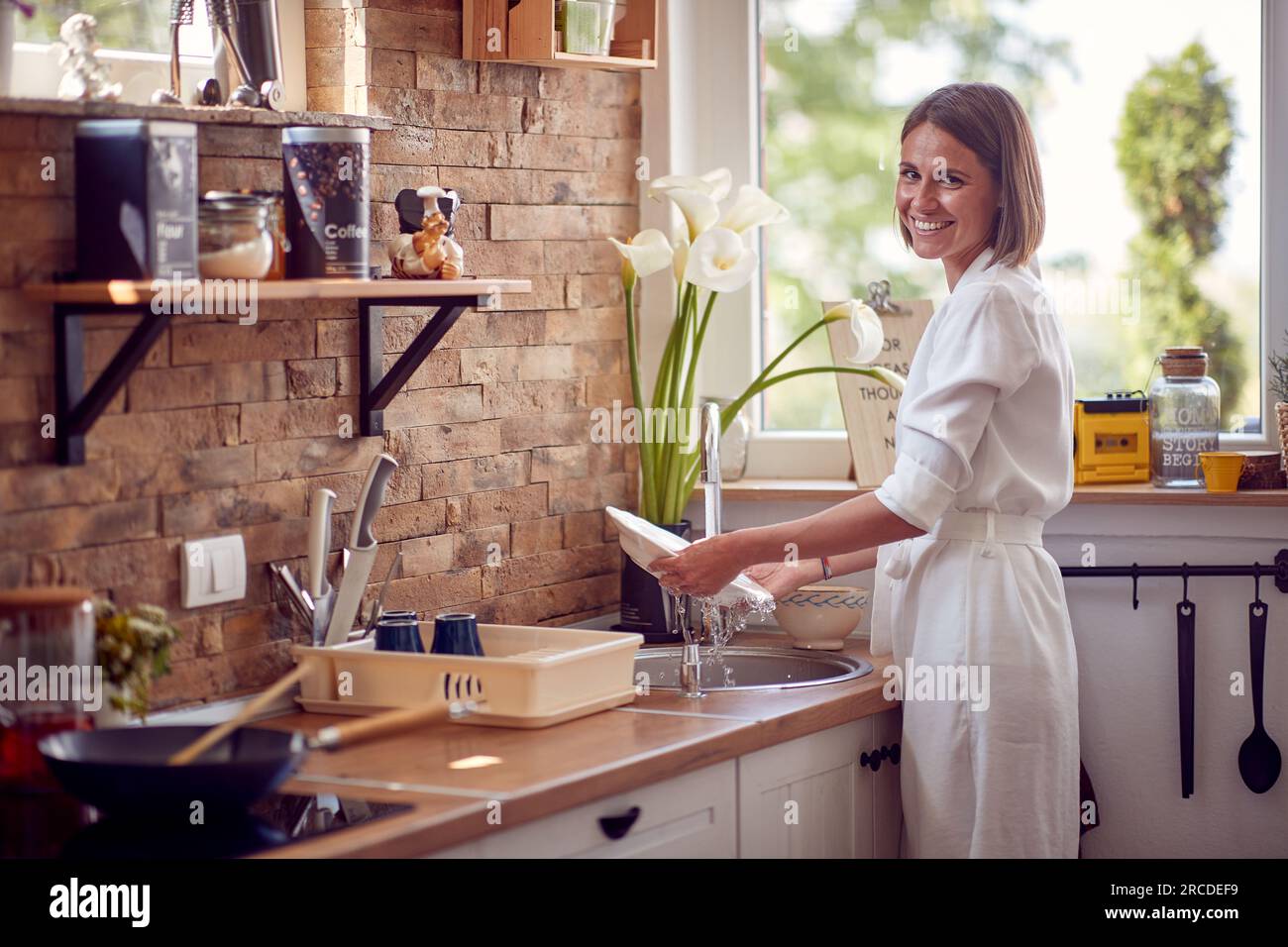 Horizontal shot of young woman washing dishes in a modern kitchen, feeling joyful. Lifestyle, home concept. Stock Photo