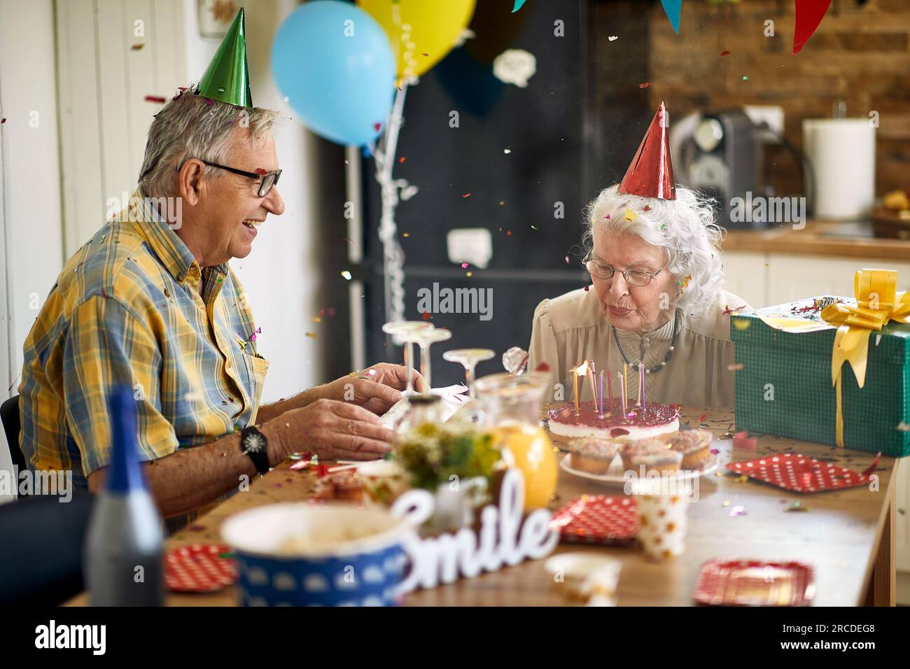 Joyful senior woman blowing candles on birthday cake, sitting at the kitchen table with her husband, celebrating her birthday. Lifestyle, senior life, Stock Photo