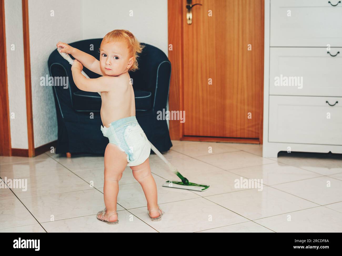 Adorable 1 year old baby boy helping with cleaning, looking back over the shoulder, wearing diaper, holding a mop Stock Photo