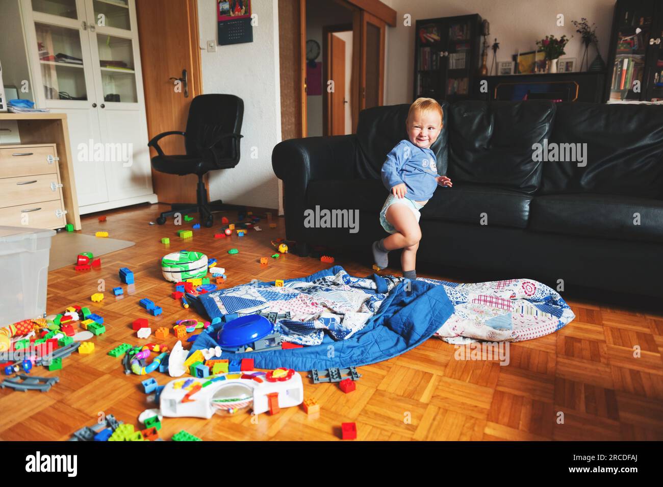 Adorable 1 year old baby boy with funny facial expression playing in a very messy living room Stock Photo