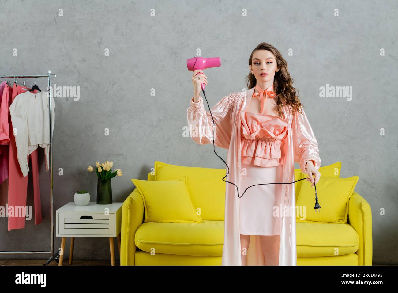 concept photography, woman acting like a doll, domestic life, housewife in pink silk robe holding hair dryer and plug, standing near yellow coach in m Stock Photo