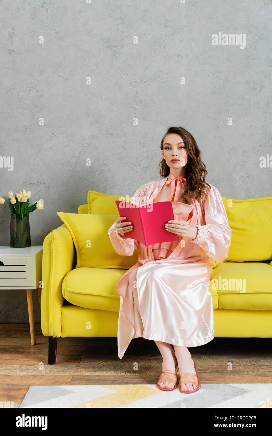 concept photography, woman with brunette wavy hair looking at camera, domestic life, attractive housewife reading book, sitting on yellow couch, comfo Stock Photo