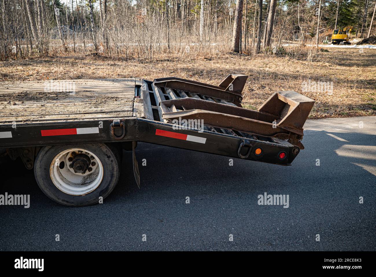 Back end of an empty flatbed trailer with raised ramps is parked on the asphalt road in a neighborhood with a house in the distance, in late afternoon Stock Photo