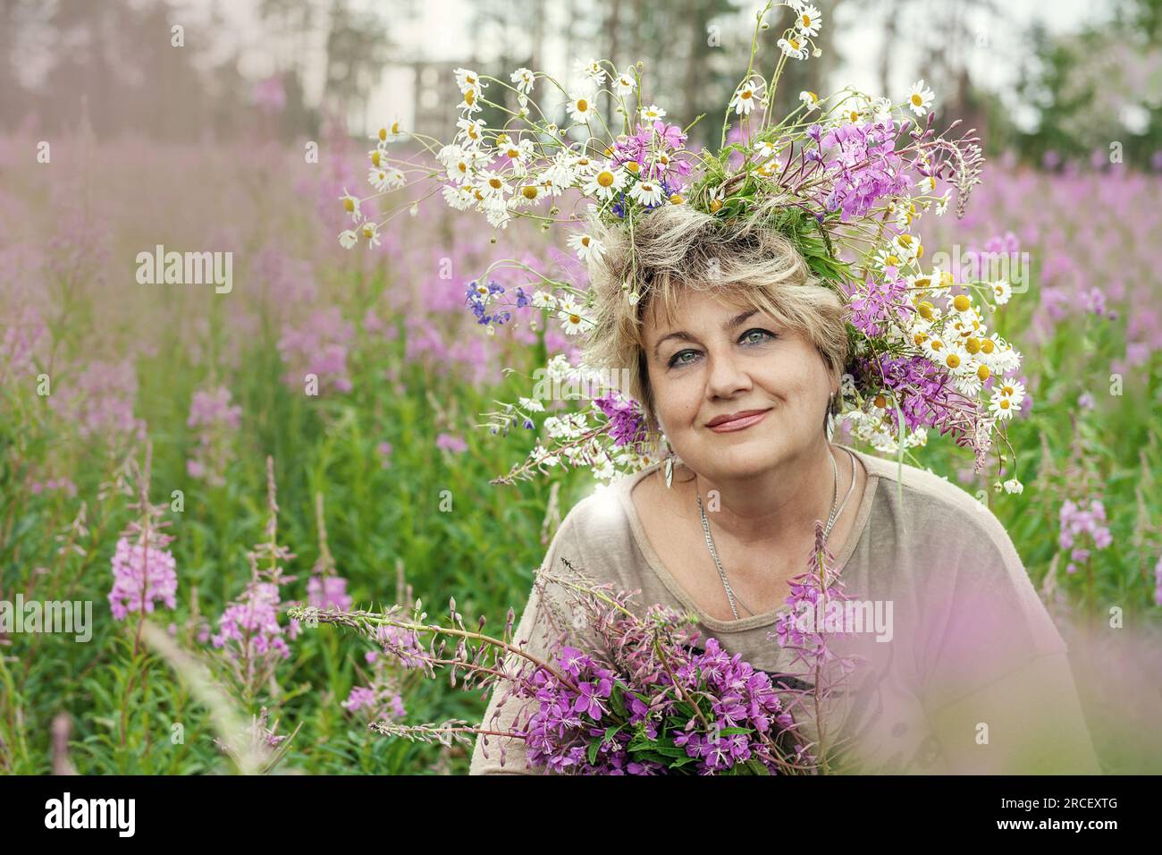 Beautiful 60s mid mature woman at nature in a a wreath of flowers in hair. Gorgeous looking old happy lady smiling. Summertime vibe. Peace and harmony Stock Photo