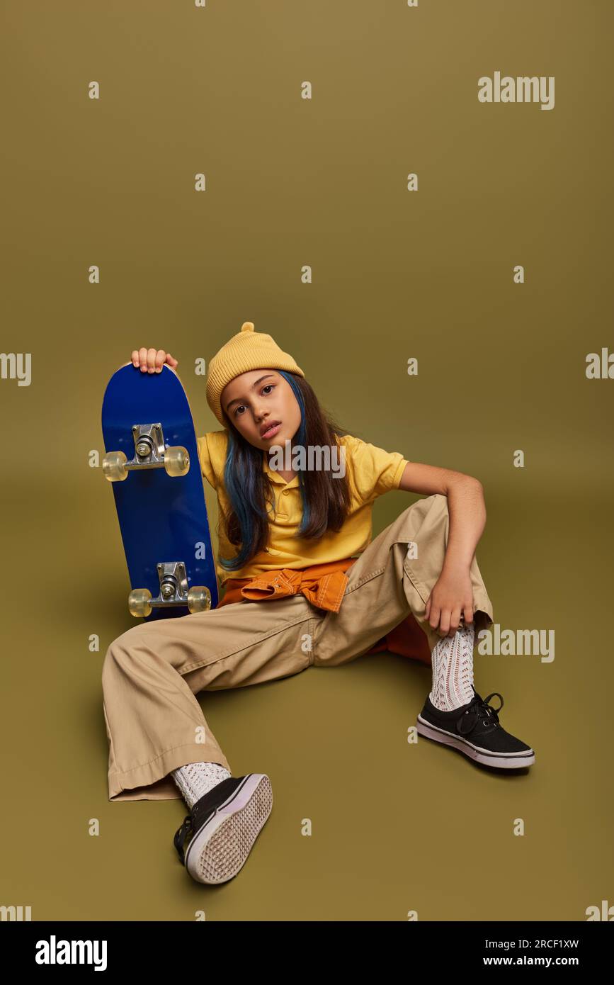 Full length of confident and fashionable preadolescent child with colored hair wearing urban clothes and hat while holding skateboard on khaki backgro Stock Photo