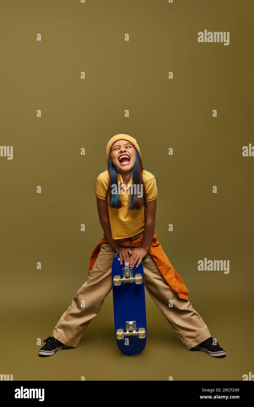 Full length of excited preteen child with colored hair wearing urban outfit and hat while grimacing and holding skateboard on khaki background, stylis Stock Photo