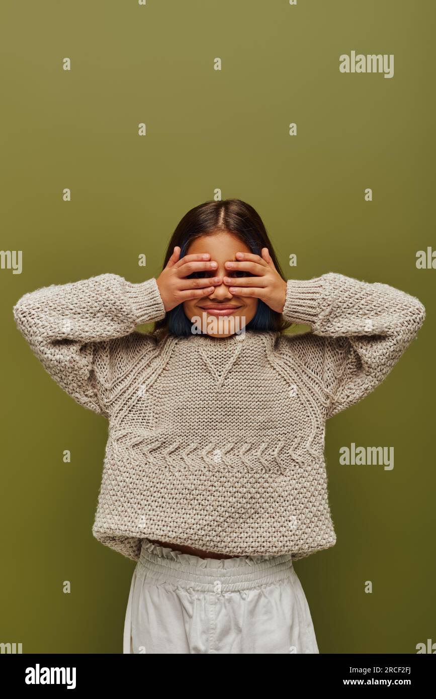 Smiling preteen child with colored hair wearing stylish knitted sweater while covering face with hands and standing isolated on green, fashion-forward Stock Photo