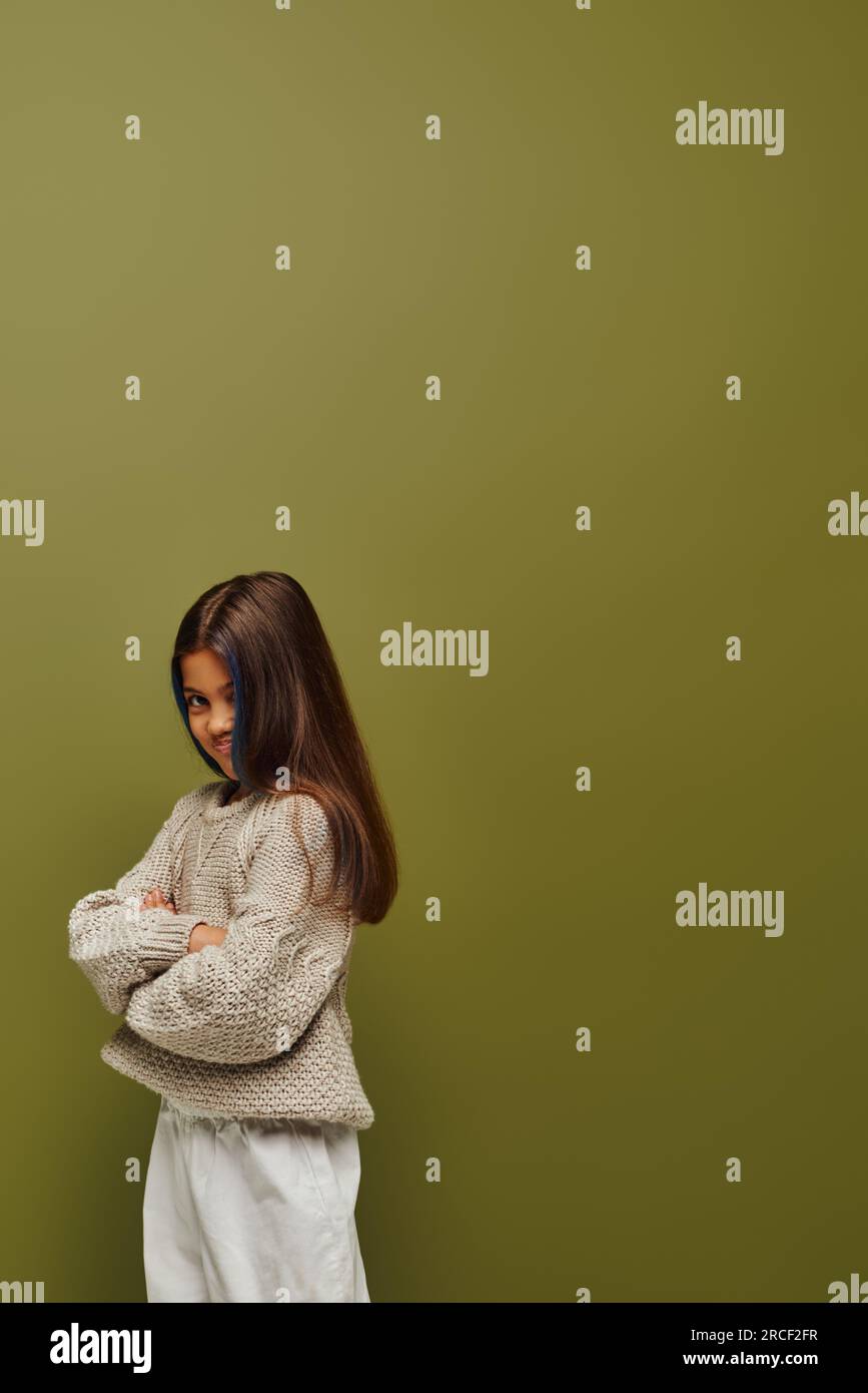 Offended and stylish preteen girl with colored hair wearing knitted sweater and crossing arms while standing and posing isolated on green, fashion-for Stock Photo