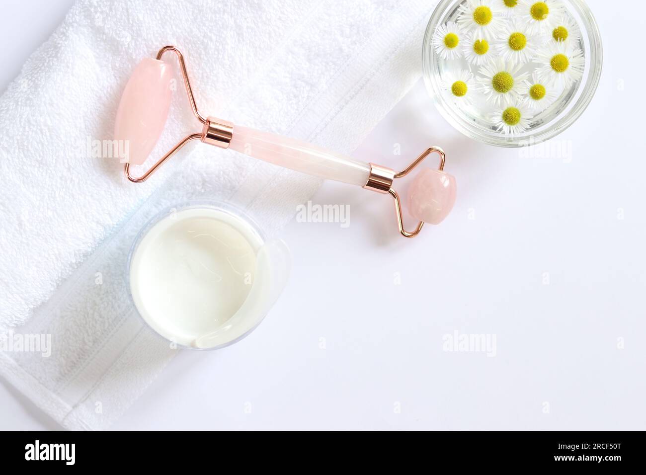Rose quartz facial roller. Facial massage roller, chamomile water, cream and white towel. Flat lay on a light background. Top view, copy space. Spa Stock Photo