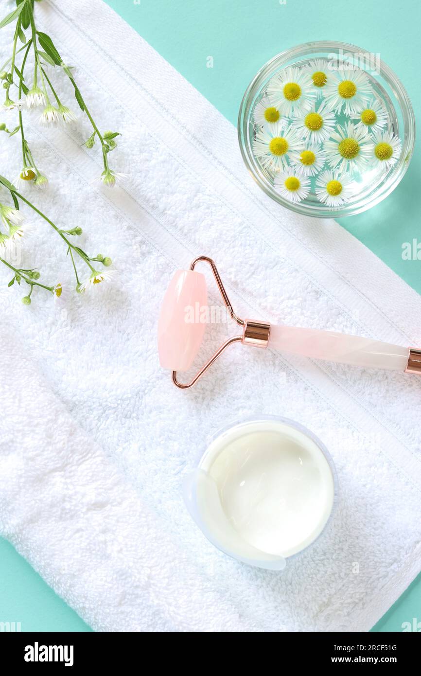 Rose quartz facial roller. Roller for cosmetic facial massage, water with chamomile flowers, cream and white towel. Flat lay, top view, copy space Stock Photo